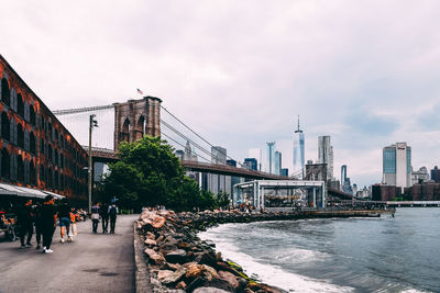 People on street by buildings and brooklyn bridge against cloudy sky