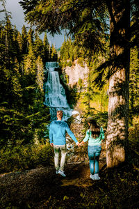 Rear view of people standing against waterfall and trees in forest