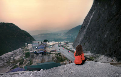 Rear view of woman sitting on rock against sky during sunset