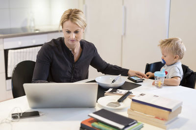 Woman working on laptop while taking care of son at dining table