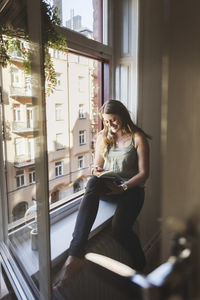 Happy woman using smart phone while holding guidebook on window sill at home