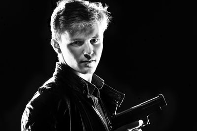 Close-up portrait of young man holding gun against black background