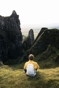 Rear view of woman sitting on rock looking at mountain against sky