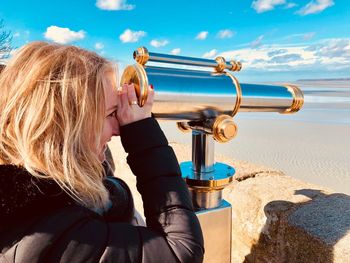 Side view of smiling woman looking through hand-held telescope at beach