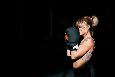 Side view portrait of young confident female athlete looking away in boxing gloves standing in defensive stance during training in dark gym