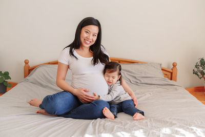 Portrait of smiling pregnant woman with daughter sitting on bed