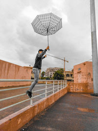 Man holding umbrella while jumping by railing against sky