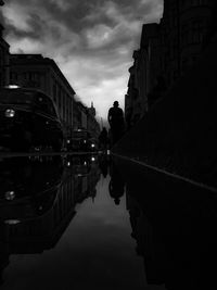 Puddle with reflection of people and cars in city against cloudy sky