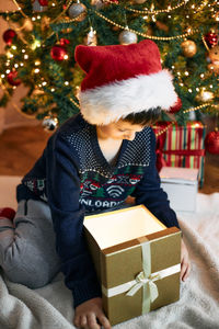 A charming boy in a santa claus hat sits next to the christmas tree and opens a box with a gift.