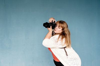 Smiling young woman photographing while standing against blue wall