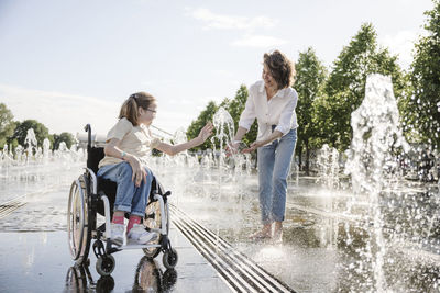 Girl in wheelchair playing along fountain water with mother at park
