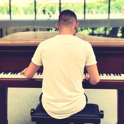 Rear view of young man playing piano while sitting indoors