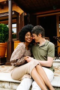 Positive multiracial couple sitting on stone stairs of porch of cottage and embracing while enjoying weekend together