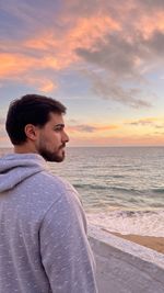 Side view of young man standing at beach against sky during sunset
