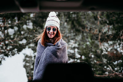 Cheerful woman wearing shawl and sunglasses standing in forest seen through car