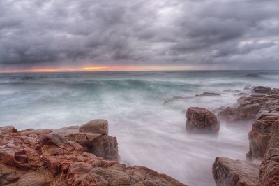 Rocks at sea shore against cloudy sky during sunset