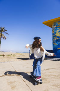Full body of active female in casual clothes riding skateboard on road along sandy beach and tall palms during training looking away