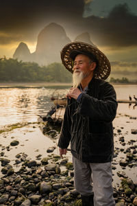 Senior man wearing hat standing by lakeshore at dusk