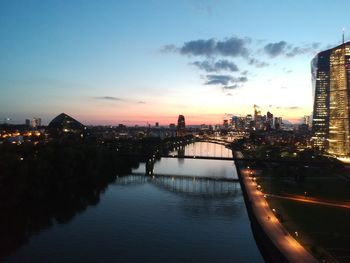 Illuminated city by river against sky during sunset