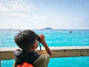 Rear view of boy looking at sea through binoculars against sky