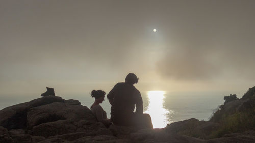 Rear view of men sitting on rock against sky