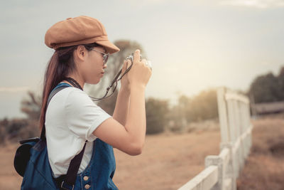 Side view of young woman photographing while standing by railing against sky