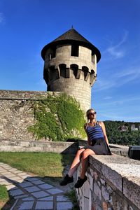 Full length of young woman sitting on retaining wall against sky