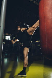 Young focused asian man training kick boxing performing strike kicks while exercising with heavy punching bag in a modern gym