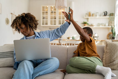 Nervous african american mother ignoring playful little boy son while working remotely on laptop