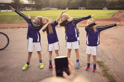 Cropped girl photographing friends performing dab dance on footpath against soccer field