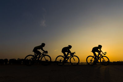 Silhouette people riding bicycles on street against sky during sunset
