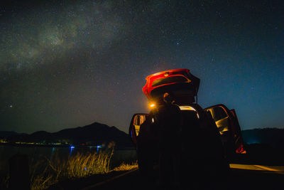 Rear view of man standing on illuminated car on road against sky at night