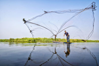 Fisherman casting fishing net while standing in lake against clear sky