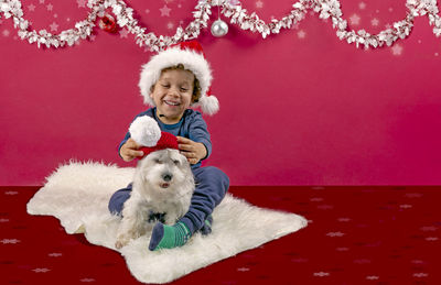  boy playing  with his little dog putting on a santa hat in a typical christmas decoration