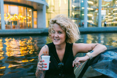 Portrait of woman holding disposable cup while sitting by pond in city