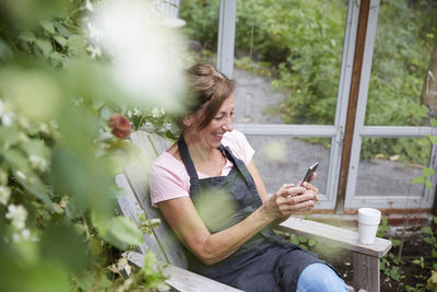 Happy female gardener using mobile phone while sitting in yard