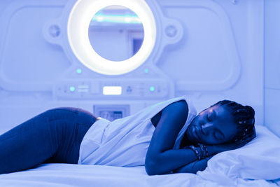 Young black woman sleeping on bed in contemporary capsule hotel with futuristic interior design and blue illumination