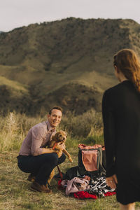 Boyfriend carrying dog while looking at girlfriend on field against mountains