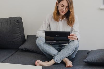 Attractive young woman working from home - female entrepreneur sitting on sofa with laptop computer and checking cell phone from comfort of home