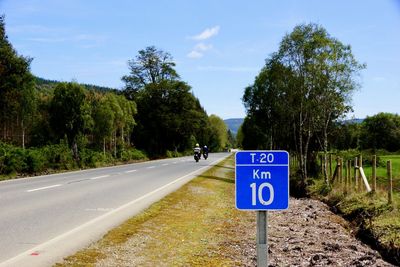 Road sign by trees against blue sky