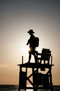 Silhouette man standing on lifeguard hut at beach against sky during sunset