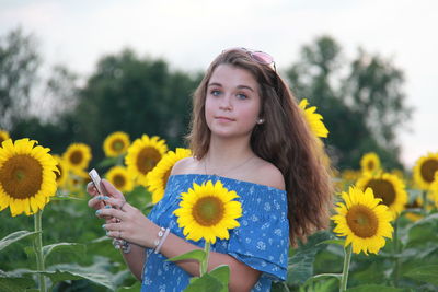 Portrait of woman holding smart phone while standing amidst sunflower field