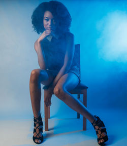 Full length portrait of confident fashion model sitting on chair in illuminated studio