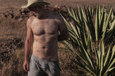 Shirtless man with cowboy hat standing by succulent plants on field