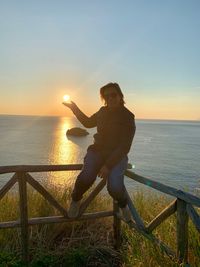 Man on railing by sea against sky during sunset