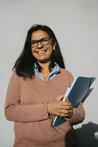 Happy mature woman with book and file standing against gray wall at university on sunny day