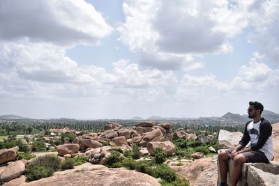 Man sitting on rock looking at mountain against sky