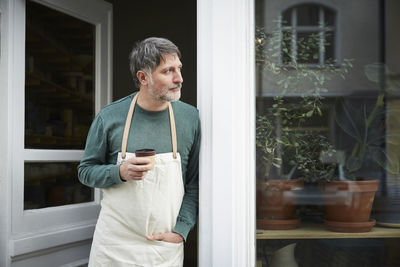 Mature male owner looking away while having coffee at entrance of art studio