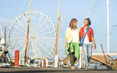 Cheerful female friends walking on pier against ferris wheel at amusement park
