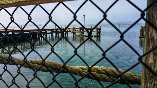 Close-up of chainlink fence and twisted rope and pier in bay water against sky